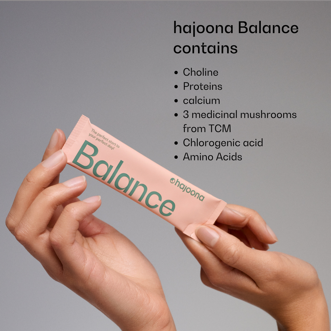 Two hands hold a pink Hajoona Balance Stick bearing green lettering. To the right is a list of its ingredients, including choline, proteins, calcium, medicinal mushrooms from Traditional Chinese Medicine (TCM), chlorogenic acid and amino acids.