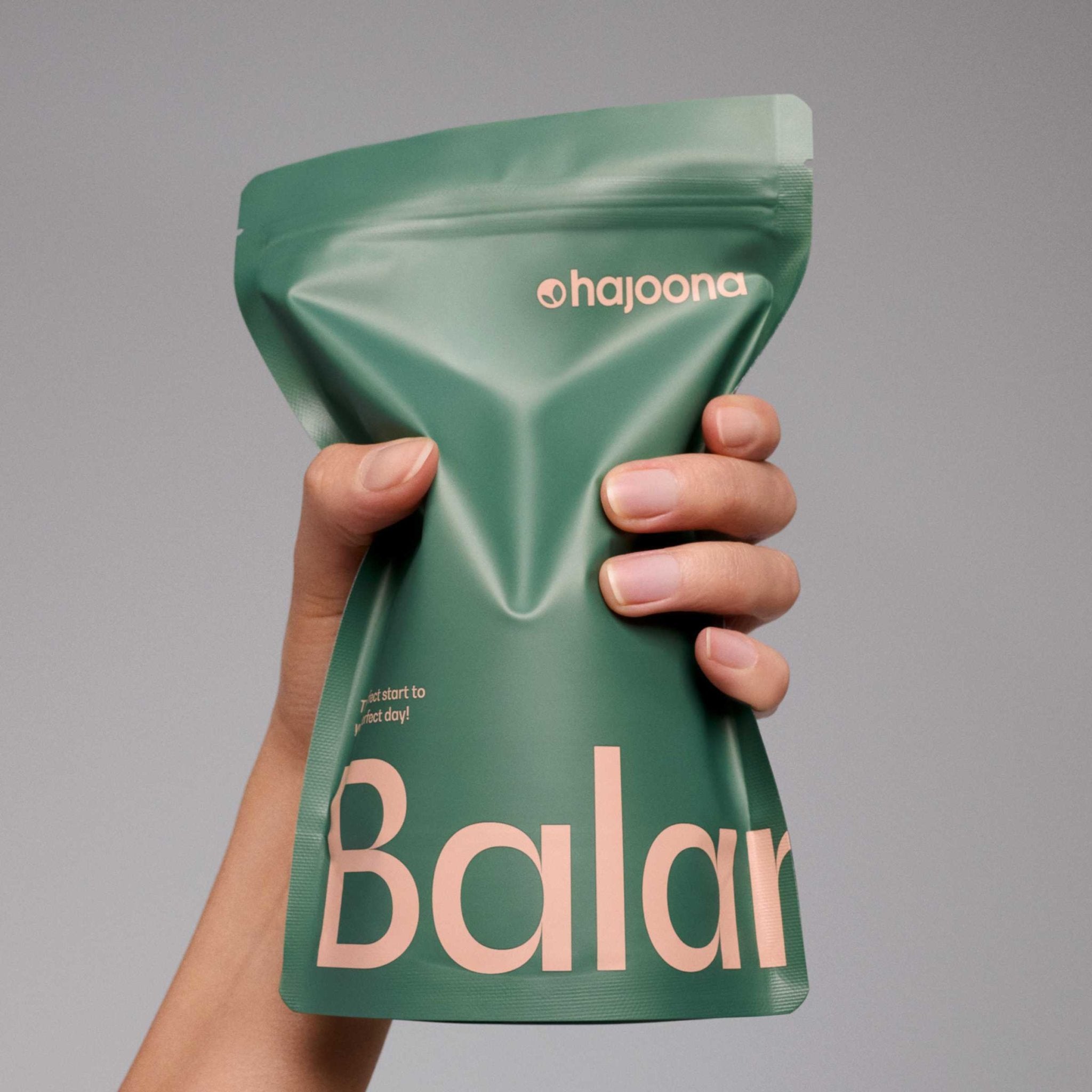 Auf dem Bild ist eine Hand zu sehen, die einen wiederverschließbaren Standbeutel von hajoona Balance hält. Die Farben Grün und Peach symbolisieren Natürlichkeit und Energie.