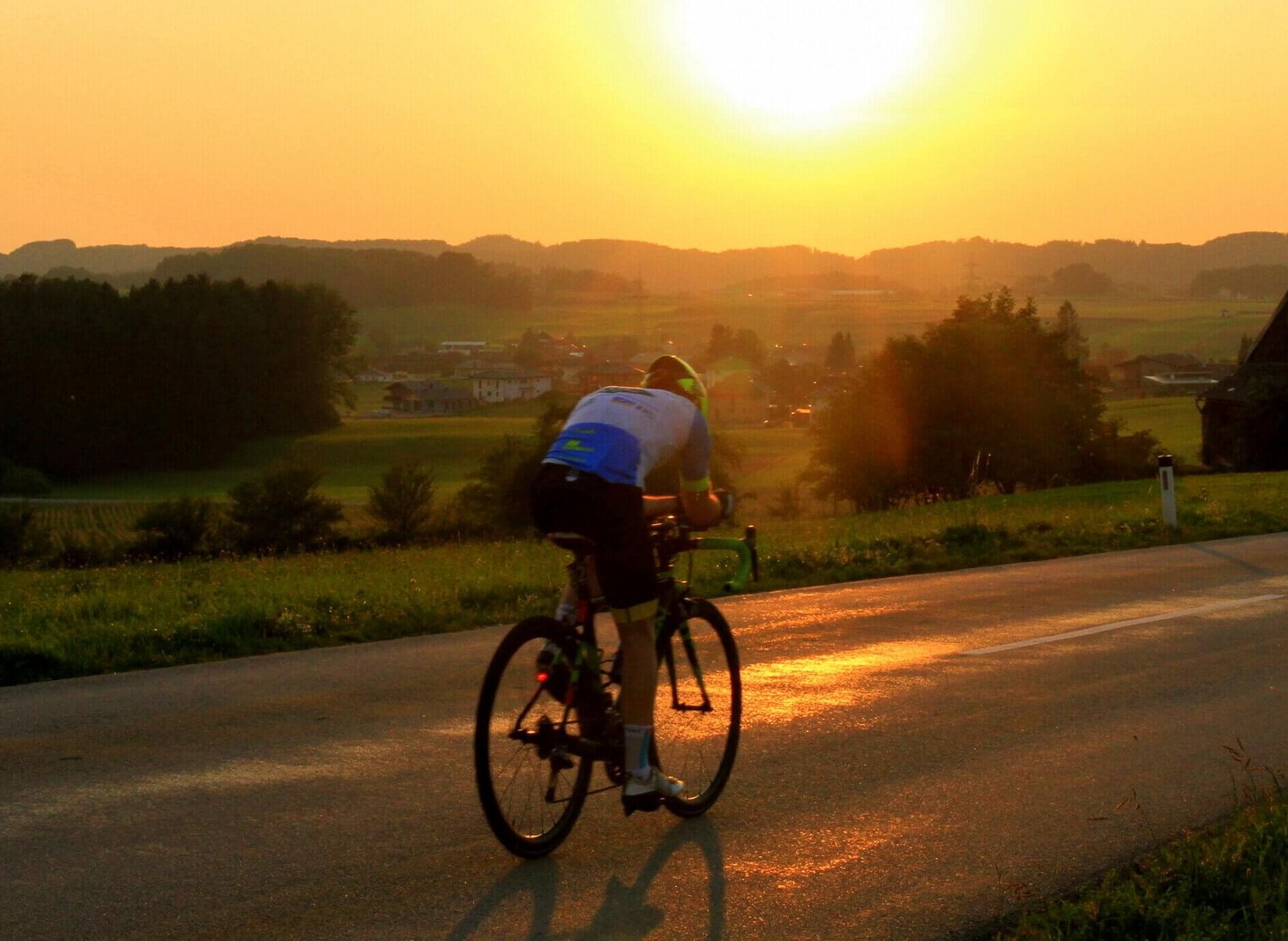 Das Bild zeigt einen Radsportler – Thomas Jaklitsch – der auf einem Rennrad eine Landstraße entlangfährt. Die Sonne steht tief am Horizont, taucht die Landschaft in goldenes Licht. Im Hintergrund sind sanfte Hügel, Bäume und ein ländliches Panorama zu sehen. Die Szene vermittelt Kraft, Ausdauer und den Moment der völligen Fokussierung. Das Bild steht im Kontext seiner Geschichte als Ultraradsportler, Mentalcoach und Speaker, der hajoona-Produkte gezielt für Energie, Erholung und Balance im Wettkampfalltag n