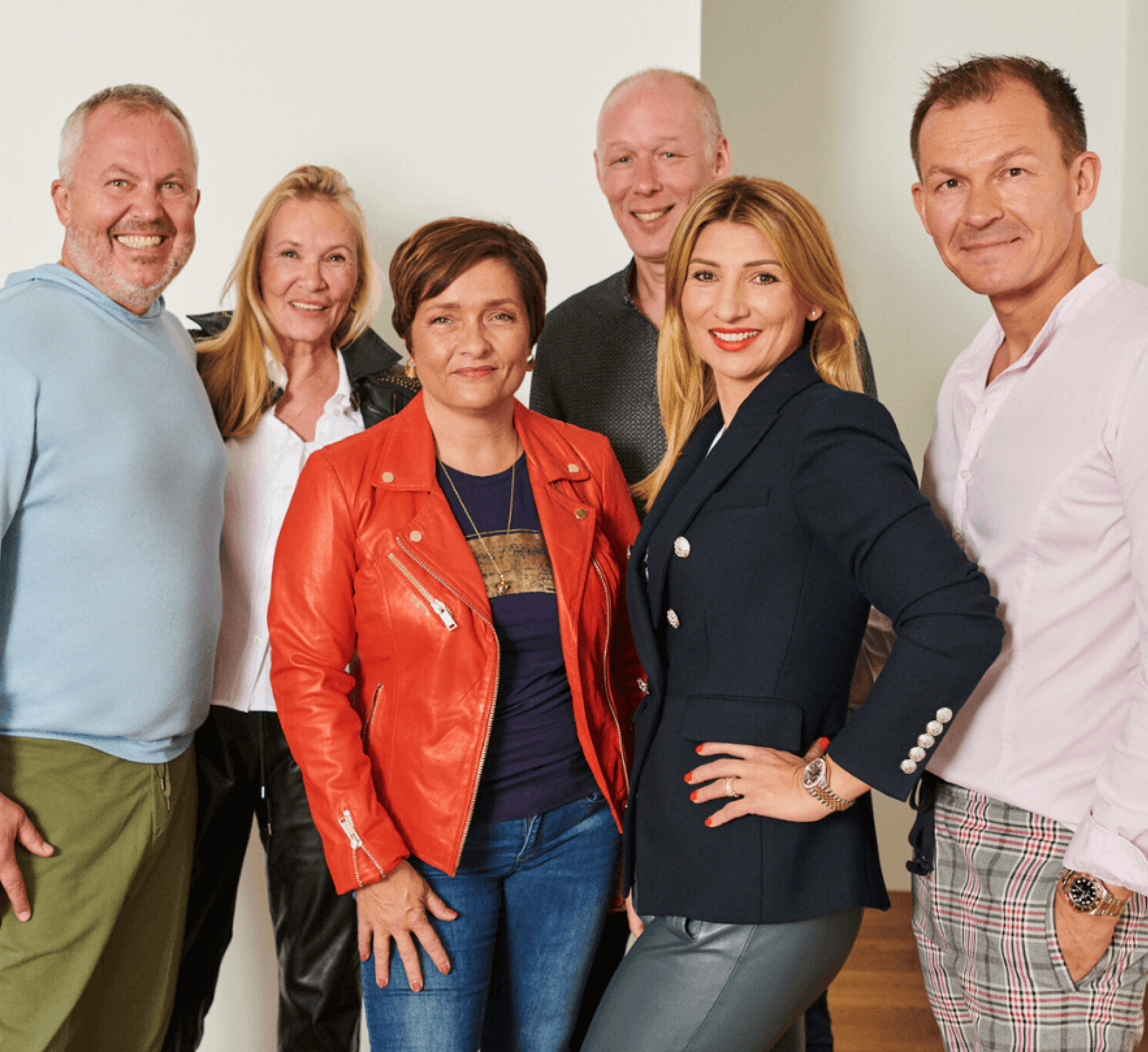 Gruppenfoto von drei Gesellschaftern der Firma hajoona – Dirk Jakob, Andrej Uschakov und Daniela Lipgens – gemeinsam mit ihren jeweiligen Partner:innen. Alle sechs Personen stehen nebeneinander vor einer hellen Wand, blicken freundlich in die Kamera und wirken entspannt und nahbar. Die Kleidung ist stilvoll und individuell, von sportlich bis elegant.