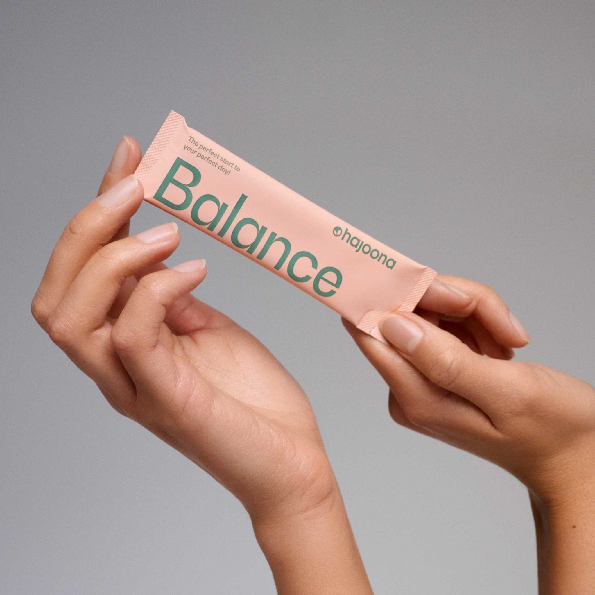 Zwei Hände halten einen rosafarbenen hajoona Sachet mit der Aufschrift ‚Balance‘ in grüner Schrift. Oberhalb steht der Claim: ‚The perfect start to your perfect day!‘. Der Hintergrund ist neutral grau und lenkt nicht vom Produkt ab.